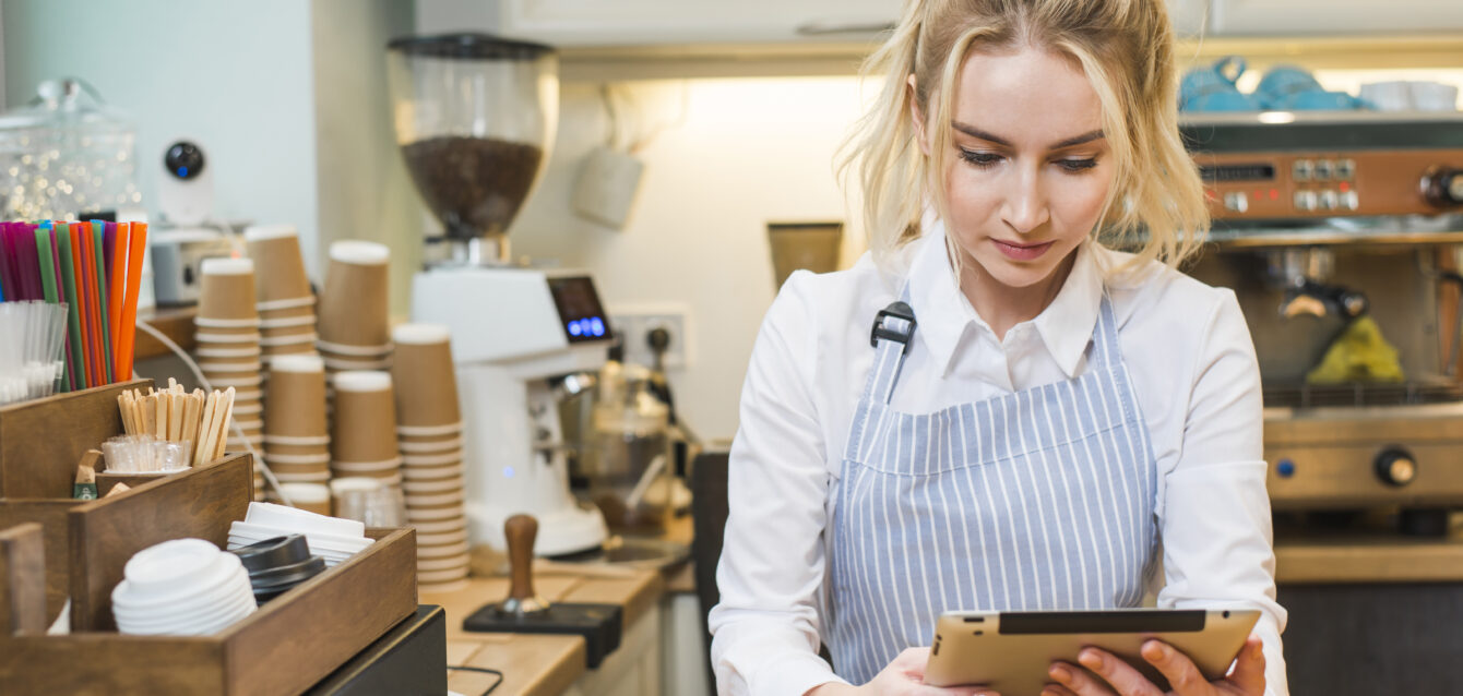 blonde-young-woman-standing-coffee-shop-counter-looking-digital-tablet