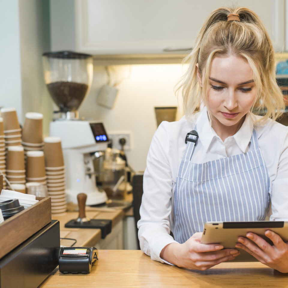 blonde-young-woman-standing-coffee-shop-counter-looking-digital-tablet